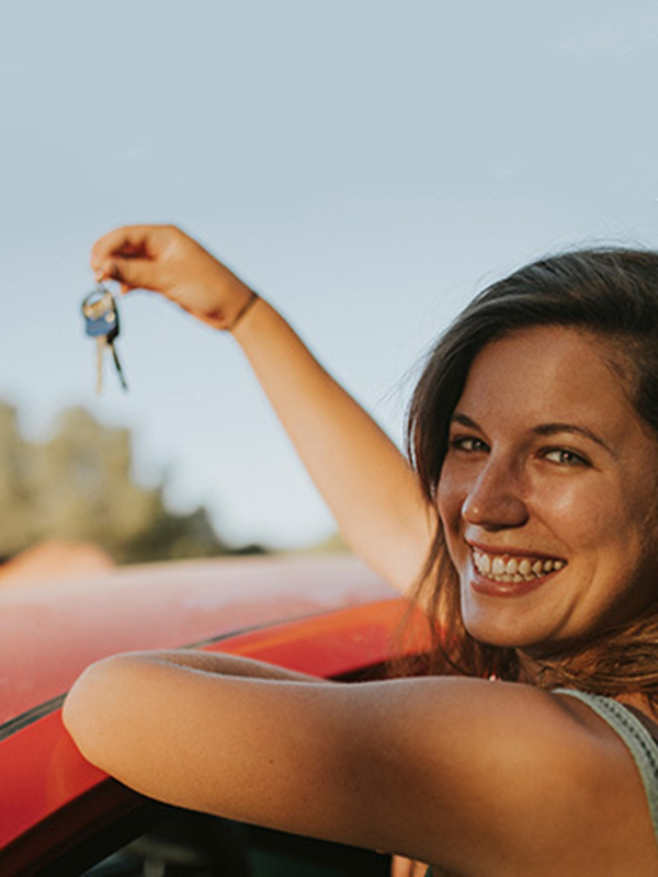 Frau lehnt lächelnd an einem Auto und hält die Schlüssel in die Luft